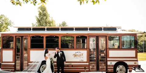 A bride and groom standing in front of a brown and white trolley bus with the Blue Bell Trolley logo.