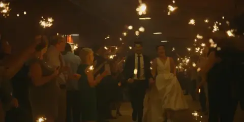 A bride and groom walking through a crowd of guests holding lit sparklers at a wedding reception.