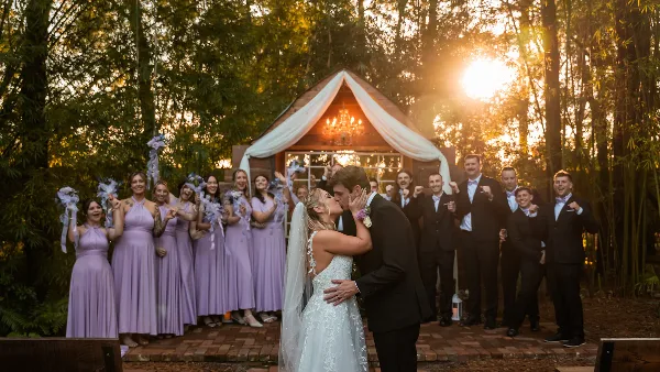 A bride and groom share a kiss in front of their wedding party at an outdoor venue during the golden hour.