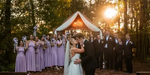 A bride and groom share a kiss in front of their wedding party at an outdoor venue during the golden hour.
