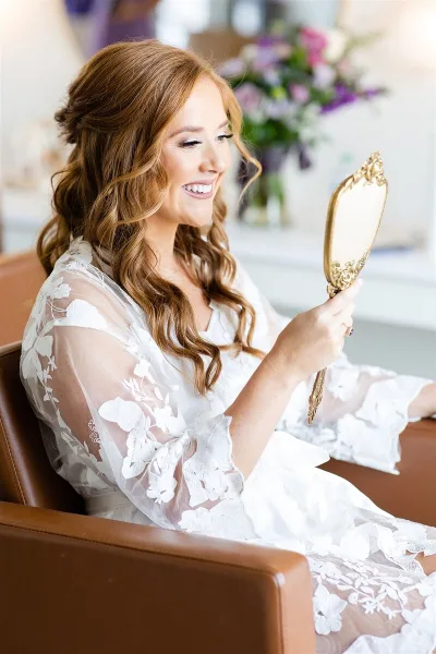 A bride with styled hair and makeup looking into a hand mirror.