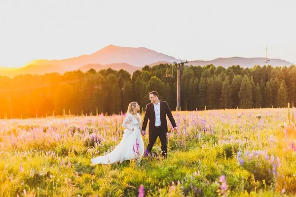 A bride and groom walking through a field of wildflowers with mountains in the background during golden hour.