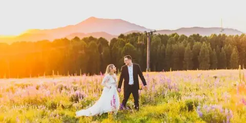 A bride and groom walking through a field of wildflowers with mountains in the background during golden hour.