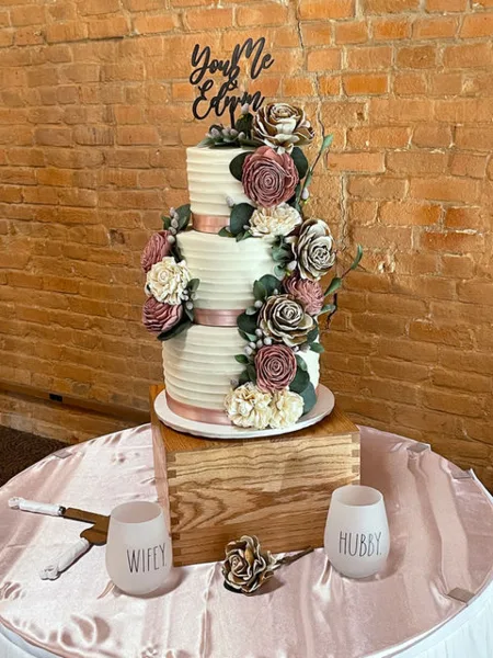 A three-tier white wedding cake decorated with pink and cream flowers, sitting on a wooden stand with 'Wife' and 'Hubby' glasses.