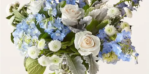 A floral arrangement with white roses, blue delphiniums, and hydrangeas in a clear glass vase.