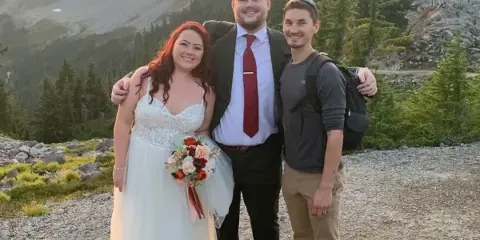 A bride and groom posing with a videographer on a mountain overlook at sunset