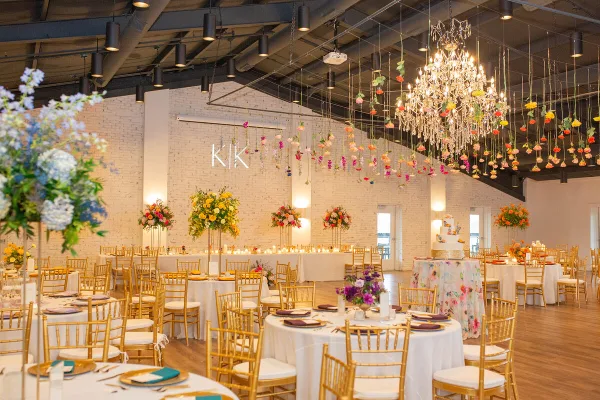 A spacious wedding reception hall featuring gold chiavari chairs, white table linens, a large floral centerpiece, and a crystal chandelier with hanging floral installations.