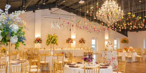 A spacious wedding reception hall featuring gold chiavari chairs, white table linens, a large floral centerpiece, and a crystal chandelier with hanging floral installations.