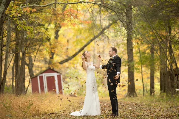 A bride and groom standing in a forest with autumn foliage, tossing leaves into the air.