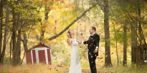 A bride and groom standing in a forest with autumn foliage, tossing leaves into the air.