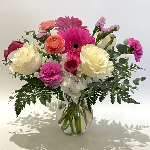 A colorful floral arrangement featuring pink gerbera daisies, white roses, and greenery in a clear glass vase.