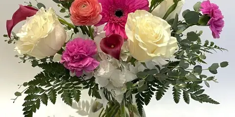 A colorful floral arrangement featuring pink gerbera daisies, white roses, and greenery in a clear glass vase.