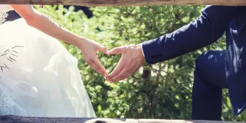 A bride and groom forming a heart shape with their hands outdoors.