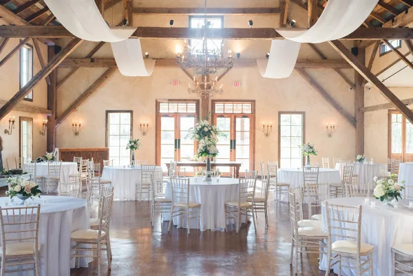 Interior of a rustic barn event venue with wooden beams, chandeliers, and round tables set for a wedding reception.
