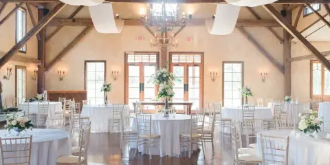 Interior of a rustic barn event venue with wooden beams, chandeliers, and round tables set for a wedding reception.