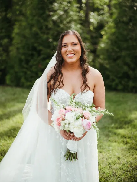 Bride in a white wedding dress and veil holding a bouquet of pink and white flowers, with styled wavy hair and makeup, standing outdoors in a grassy area.