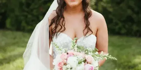 Bride in a white wedding dress and veil holding a bouquet of pink and white flowers, with styled wavy hair and makeup, standing outdoors in a grassy area.
