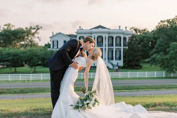 A bride and groom share a kiss in a dip pose on a lawn in front of a large white plantation-style house.