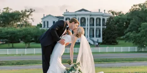 A bride and groom share a kiss in a dip pose on a lawn in front of a large white plantation-style house.