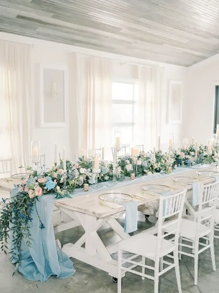 A long white wooden table set for a wedding reception with floral centerpieces, candles, and white Chiavari chairs in a bright room.