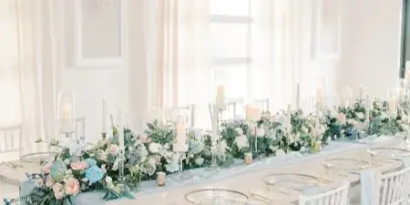 A long white wooden table set for a wedding reception with floral centerpieces, candles, and white Chiavari chairs in a bright room.
