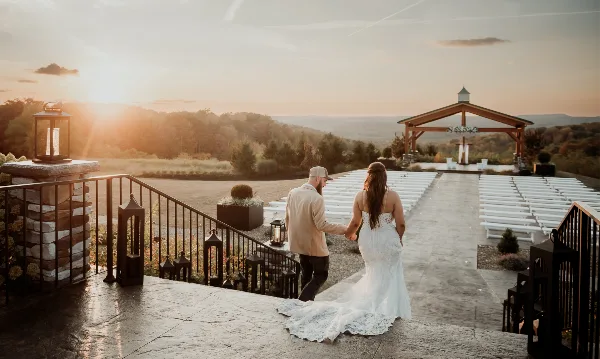 A bride and groom holding hands on a stone terrace overlooking a wedding ceremony setup with white chairs and a wooden arch at sunset.