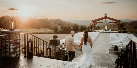 A bride and groom holding hands on a stone terrace overlooking a wedding ceremony setup with white chairs and a wooden arch at sunset.
