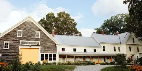 Exterior view of a rustic wooden barn with a yellow door and an adjacent white farmhouse building.