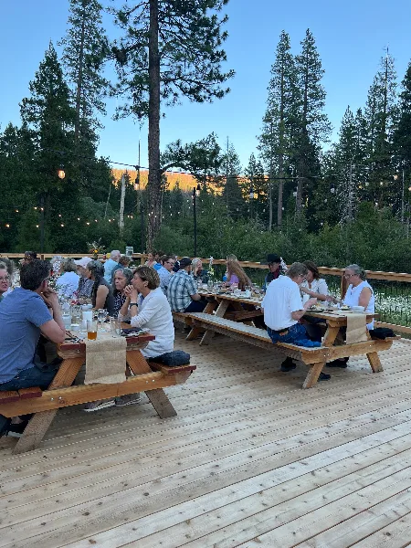 Guests seated at long wooden tables on a deck for a meal, surrounded by tall pine trees and string lights.