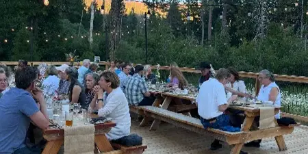 Guests seated at long wooden tables on a deck for a meal, surrounded by tall pine trees and string lights.