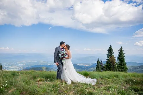 A bride and groom kissing on a grassy mountain summit with a view of the valley below.