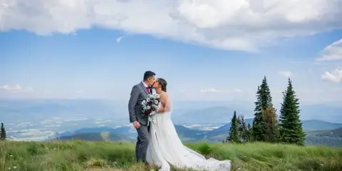 A bride and groom kissing on a grassy mountain summit with a view of the valley below.