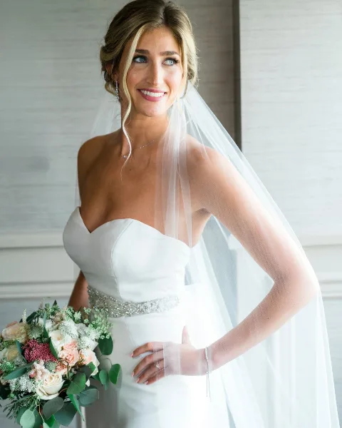A professional portrait of a smiling bride in a white strapless gown and veil, holding a bouquet of flowers.
