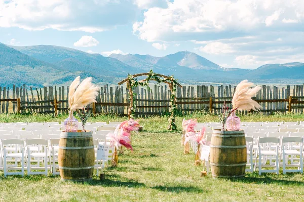 Outdoor wedding ceremony setup with white chairs and a floral arch in a grassy field with a mountain backdrop.