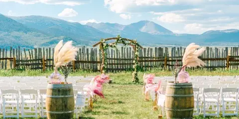 Outdoor wedding ceremony setup with white chairs and a floral arch in a grassy field with a mountain backdrop.
