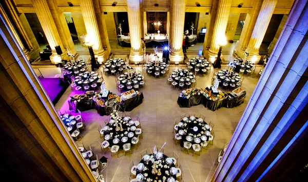 A grand banquet hall with tall columns, set with numerous round dining tables and buffet stations for a large event.