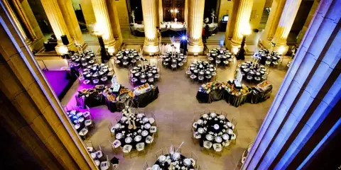 A grand banquet hall with tall columns, set with numerous round dining tables and buffet stations for a large event.