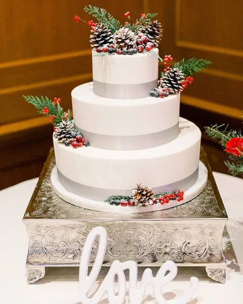 A three-tier white wedding cake decorated with pinecones, red berries, and greenery, sitting on a silver pedestal stand.