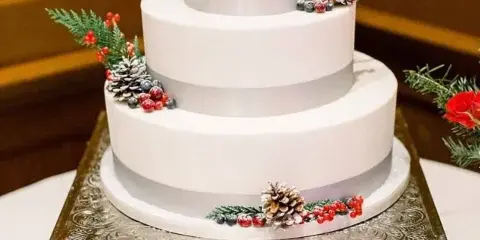 A three-tier white wedding cake decorated with pinecones, red berries, and greenery, sitting on a silver pedestal stand.