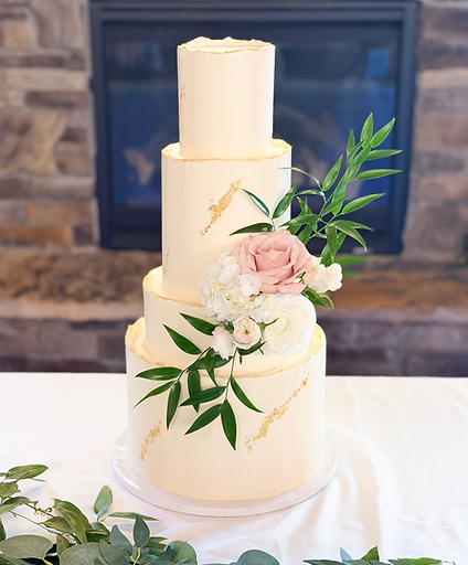 A three-tiered white wedding cake with fresh floral decorations and gold leaf accents on a table.