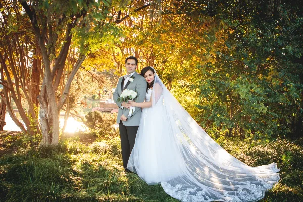 A bride and groom posing together outdoors in a wooded area with golden sunlight filtering through the trees.