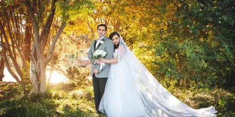 A bride and groom posing together outdoors in a wooded area with golden sunlight filtering through the trees.
