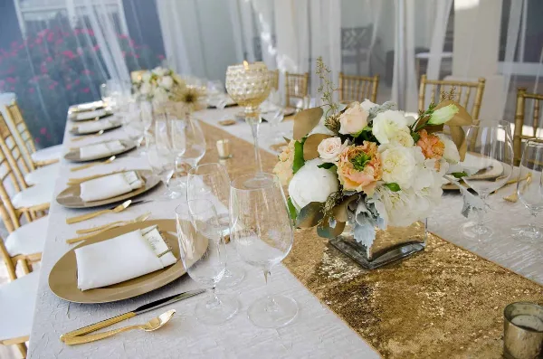 A close-up view of a formal event table setting featuring gold Chiavari chairs, white table linens, and a gold sequin runner.