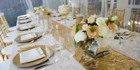 A close-up view of a formal event table setting featuring gold Chiavari chairs, white table linens, and a gold sequin runner.