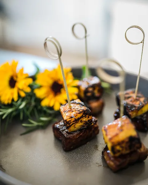 A close-up of a plated appetizer featuring a skewered cube of food, garnished with yellow flowers and rosemary.