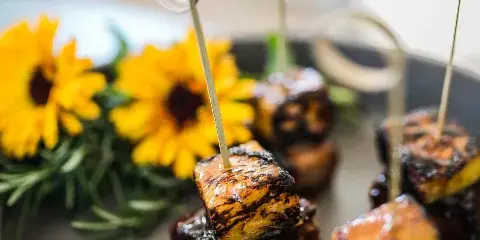 A close-up of a plated appetizer featuring a skewered cube of food, garnished with yellow flowers and rosemary.