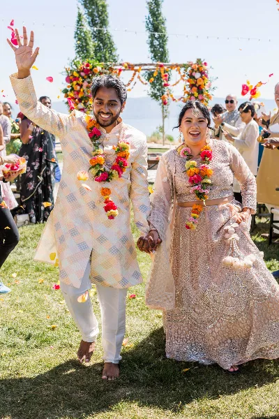 A smiling bride and groom in traditional Indian wedding attire walking hand-in-hand while guests throw flower petals around them.