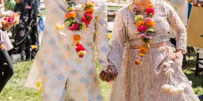 A smiling bride and groom in traditional Indian wedding attire walking hand-in-hand while guests throw flower petals around them.