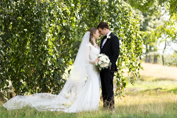 A bride and groom standing close together in a grassy field with a large leafy tree in the background.