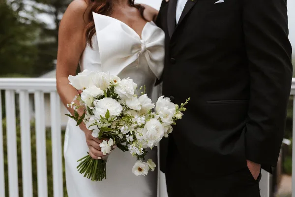A bride in a white gown holding a white floral bouquet standing next to a groom in a black suit.